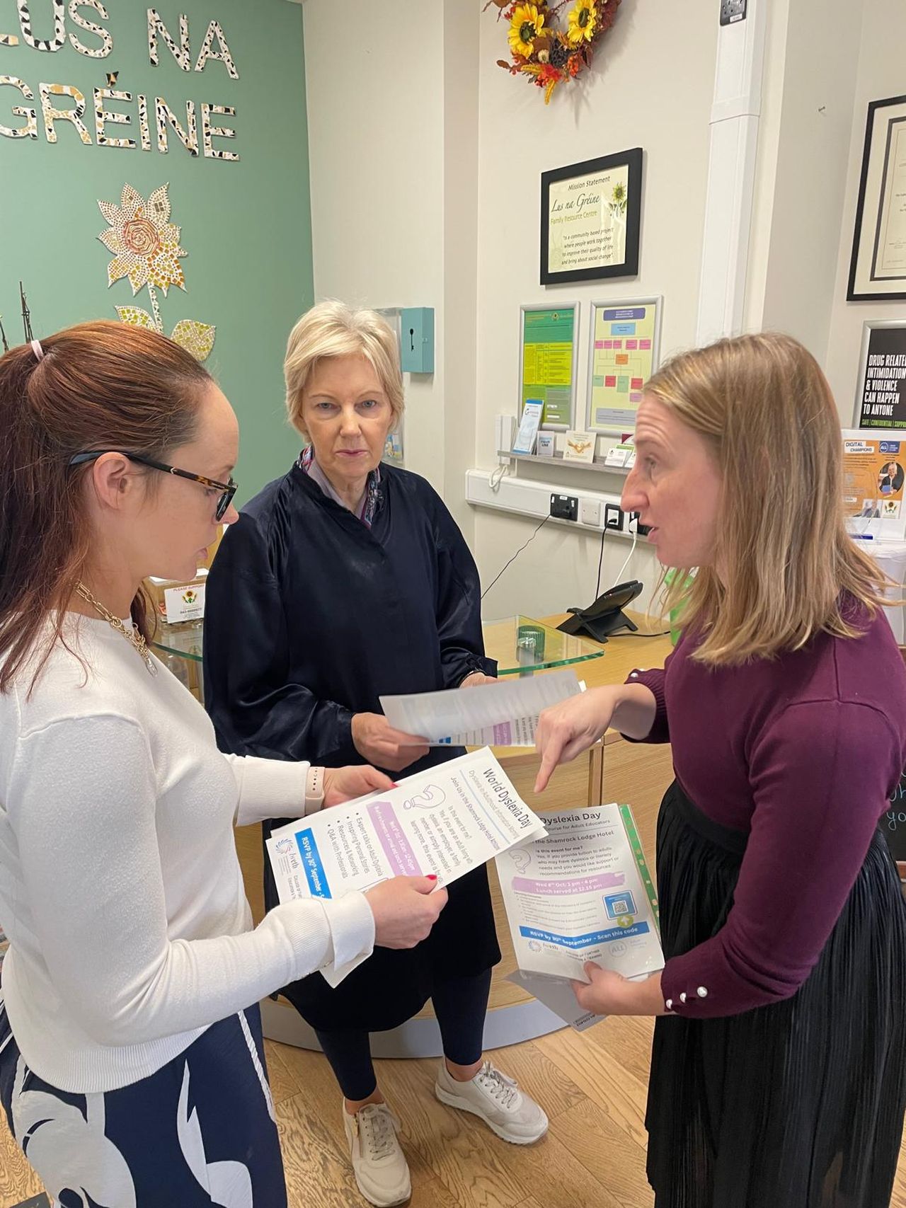Three women are in discussion, holding informational flyers in an office decorated with sunflowers and motivational posters.